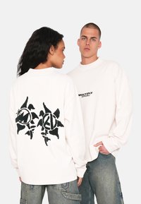 Weiße Langarm-T-Shirts aus Baumwolle mit einem schwarzen Grafikdesign auf dem Rücken und einem kleinen Logo auf der Vorderseite. Legere Passform.
