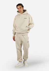 Beige bomull hettegenser med frontlomme og svart logo, paret med matchende joggebukser med sidelommer og elastiske mansjetter. Hvite sneakers.