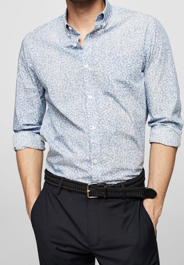 Homme portant une chemise à boutons bleu clair à motif floral avec manches retroussées et ceinture tissée noire sur un pantalon foncé.