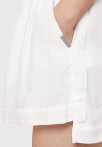 Tunic bianca realizzata in un tessuto leggero e textured; presenta una tasca laterale e un'etichetta argentata con il marchio vicino all'orlo. Finitura liscia e vestibilità comoda.