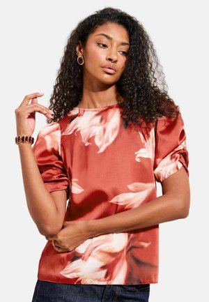 Frau mit lockigen Haaren, die eine rote Satinbluse mit Blumenmuster und passenden Schmuck trägt, posiert mit einem erhobenen Arm und schaut seitlich.