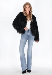 Eine lächelnde Frau steht in einer schwarzen strukturierten Jacke, einem weißen Oberteil, hellblauen high-waisted Jeans, schwarzen Schuhen und rosa Quastenohrringen.