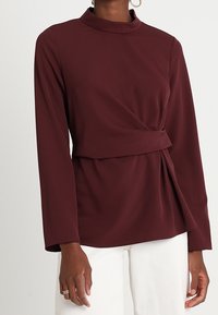 Blusa bordeaux a maniche lunghe con collo alto, design avvolgente e dettaglio drappeggiato laterale, realizzata in tessuto liscio. Indossata con pantaloni bianchi.