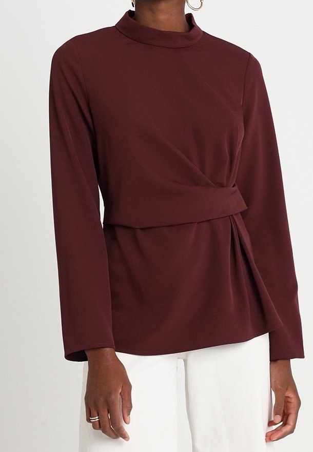 Blusa bordeaux a maniche lunghe con collo alto, design avvolgente e dettaglio drappeggiato laterale, realizzata in tessuto liscio. Indossata con pantaloni bianchi.