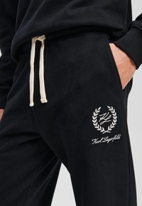 Pantaloni sport de culoare neagră, confecționați dintr-un material moale, cu un șnur alb, buzunar lateral și un logo brodat cu accente de laur pe coapsă.