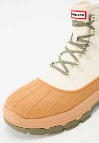 Botine impermeabile Hunter cu vârful din cauciuc bej, partea superioară din pânză crem, șireturi verzi și talpă texturată pentru aderență.