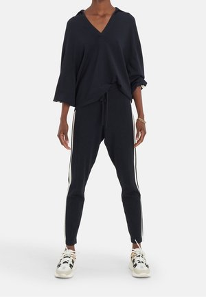 Personne portant un haut noir ample à col en V, un jogging noir avec des bandes blanches sur les côtés, des baskets blanches et noires, et un bracelet en or au poignet droit.