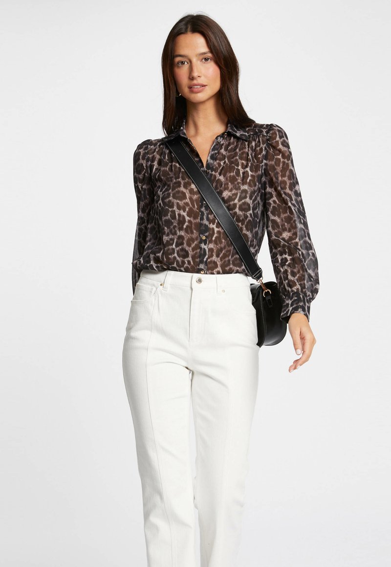 Blouse en imprimé léopard marron et noir semi-transparent, avec des manches longues, un devant boutonné et une silhouette cintrée, assortie à un pantalon blanc taille haute.