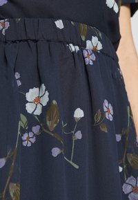 Šaty s námořnickou modrou elastickou páskou včetně bílého a fialového květinového vzoru, ukazující detailní texturu látky a oblast pasu na osobě.