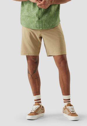 Mann trägt beige Shorts, ein grünes gemustertes Hemd, weiße Socken mit braunen Streifen und beige Sneakers, steht vor einem grauen Hintergrund.