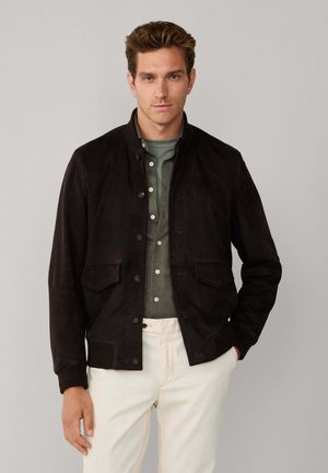 Hombre con chaqueta negra abotonada sobre camisa verde y pantalones blancos, de pie frente a un fondo liso gris claro.