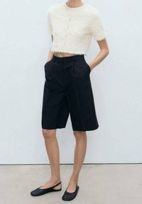 Haut crème texturé et raccourci avec manches courtes ; shorts noirs taille haute avec plis et poches latérales ; chaussures plates noires.