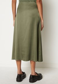 Falda midi verde oliva de tela suave, con un diseño fluido y un dobladillo ligeramente acampanado. Combinada con zapatos de plataforma negros y gruesos.