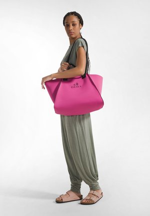 Pink tote bag with black accents, made of soft material, featuring a rounded shape and logo. Model wears a light green outfit and sandals.