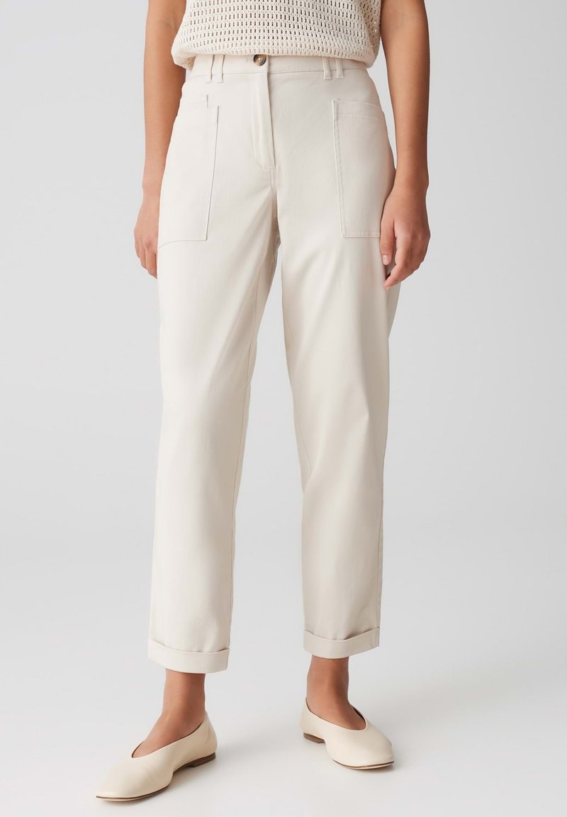 Pantalons couleur crème avec une coupe décontractée, des poches plaquées à l'avant, des ourlets à revers et une texture de tissu légère. Associés à des chaussures légères et arrondies.