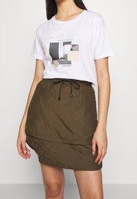 Vit t-shirt med abstrakt grafik i nyanser av grått och beige, kombinerad med en brun, vadderad, dubbel-lager kjol med snörning.