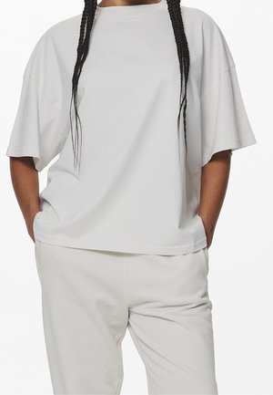 Person trägt locker sitzendes weißes T-Shirt und passende weiße Hose mit den Händen in den Taschen und langem geflochtenem Haar.