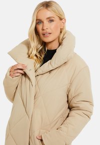 Beige polstret frakke med en stor krave, diamantquiltet mønster og trykknaplukning. Har sidelommer og en afslappet pasform.