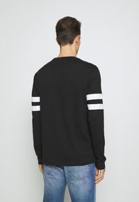 Svart långärmad t-shirt med vita ränder på ärmarna, tillverkad av mjukt tyg. Bakifrån visas en rund halsringning och en avslappnad passform.