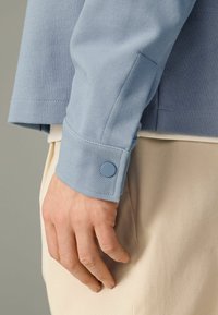Tela texturizada de color azul claro de la manga de una camisa con un puño redondeado, que presenta un botón a juego. Un tejido beige aparece en el fondo.