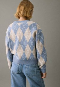 Maglione a motivi argyle blu e beige realizzato in tessuto a maglia, con polsini e orlo a coste, indossato con jeans blu chiaro.
