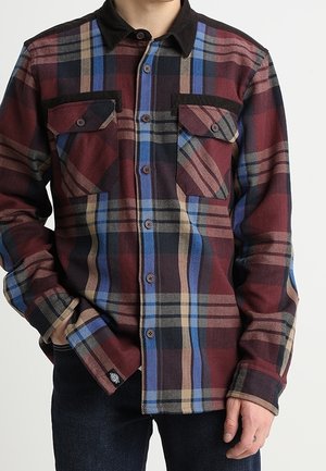 Uomo che indossa una camicia di flanella a maniche lunghe con motivo a quadri nei colori rosso, blu, nero e beige, con due tasche sul petto chiuse da bottoni.
