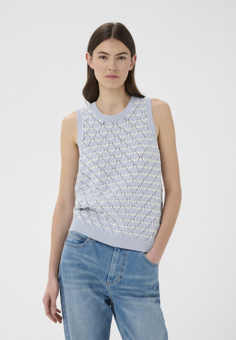 Femme portant un haut sans manches en maille violet clair à motif losange et un jean bleu clair, debout avec une main dans la poche.