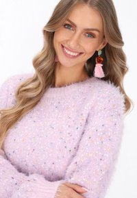 Jasnofioletowy, puszysty sweter o teksturowym wykończeniu, z kolorowymi drobinkami i okrągłym dekoltem. Modelka nosi różowe kolczyki z frędzlami.