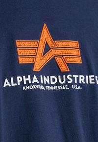 Marineblå bomull T-skjorte med en oransje strukturert logo og hvit tekst som sier "ALPHA INDUSTRIES" med stedsdetaljer under.
