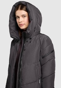 Geacă matlasată gri cu glugă mare, design matlasat, închidere cu fermoar frontală și buzunar lateral cu accent negru.