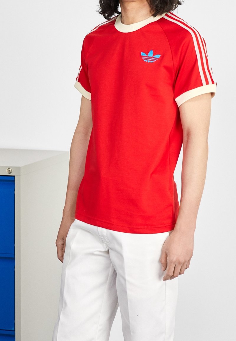 Červené bavlněné tričko s krémovým žebrovaným límečkem a rukávy. Obsahuje modrou vyšitou logo a bílé pruhy na ramenou. Kombinováno s bílými kalhotami.