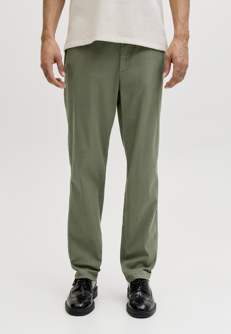 Hombre que lleva pantalones verde oliva de pierna recta, zapatos de vestir negros y una camisa texturizada de color claro contra un fondo liso.