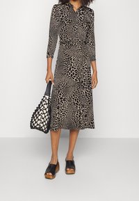 Leopardmuster-Kleid mit langen Ärmeln, Taillengürtel und mittlerer Länge. Kombiniert mit einer schwarzen Netz-Handtasche und schwarzen Plateau-Holzschuhen.
