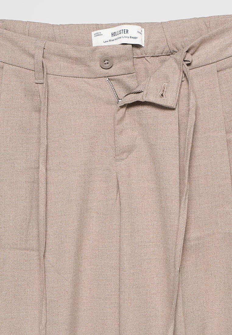 Beige, weit geschnittene Baggy-Hosen mit niedrigem Bund, teilweise offenem Reißverschluss, Knopfverschluss, Gürtelschlaufen und sichtbarem Hollister-Logo im Saum.