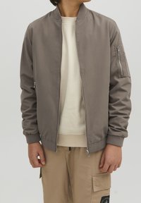Persona vestida con una cazadora bomber en tono topo con cremallera frontal, sudadera beige y pantalones cargo beige con un parche en el muslo izquierdo.