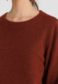 Rostfarbener Strickpullover mit Rundhalsausschnitt und gerippten Bündchen. Weiche Textur mit einer leicht lockeren Passform, die ein dezentes Muster zur Geltung bringt.