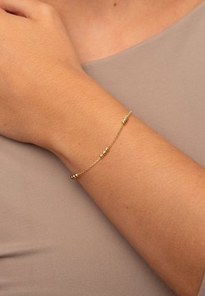 Bracciale d'oro con un delicato design a catena, caratterizzato da piccole perle rotonde distanziate uniformemente, poggiate su un polso abbronzato su uno sfondo neutro.