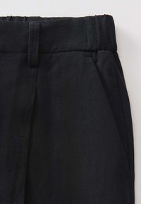 Gros plan sur un pantalon en lin noir montrant une taille élastique, une boucle de ceinture et une poche latérale sur fond blanc.