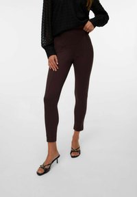 Donkerbruine, strakke broek met een gladde textuur, hoge taille en naaddetails, gecombineerd met zwarte open hakken. Zwarte, gestructureerde blouse.
