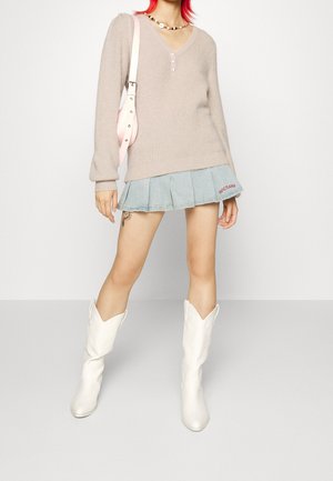 Suéter acanalado de color beige claro con tres botones, combinado con una falda de mezclilla plisada que presenta bordado rojo "EDGETEANS" y botas de cowboy blancas.