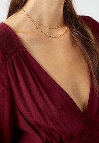 Collares de oro en capas combinados con una blusa burdeos profunda que presenta un escote en V y detalles fruncidos en los hombros. Textura suave en general.