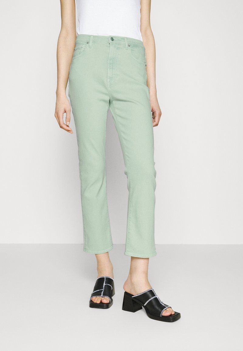 Jeans en denim vert clair à coupe droite, cinq poches, et boutons en métal, accompagnés de sandales noires à talons blocs.
