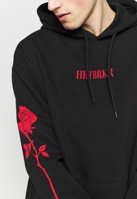 Jeune personne portant un sweat à capuche noir avec le texte rouge "HRTBRKR" sur la poitrine et un motif de rose rouge le long d'une manche, regardant vers le bas et sur le côté.