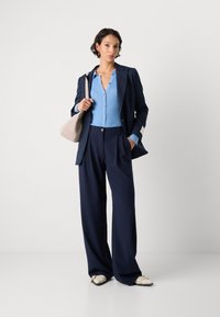 Marineblauer Nadelstreifen-Blazer über einem hellblauen gerippten Cardigan, kombiniert mit weit geschnittenen marineblauen Hosen und einer beigen Tasche. Mit spitzen, genagelten Schuhen.