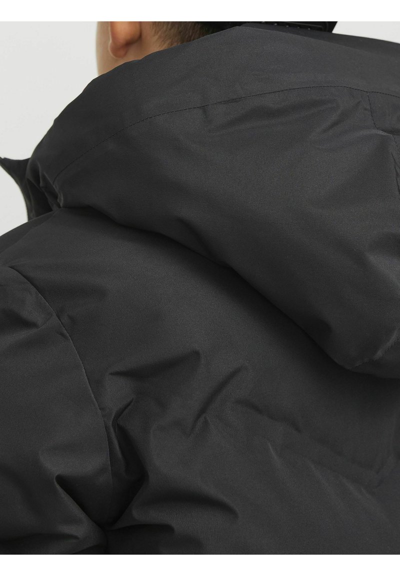 Schwarze Steppjacke mit passender Kapuze, aus glattem, mattem Stoff mit sichtbaren Nähten. Fokus auf der Rückansicht und der Textur.