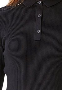 Gros plan d'une personne portant un polo noir côtelé à manches longues avec trois boutons noirs sur la patte de boutonnage.