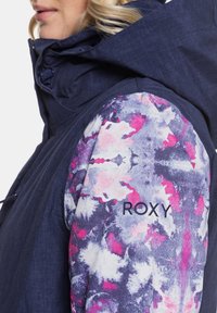 Marineblaue Jacke mit einem gemusterten Ärmel in Pink- und Violetttönen; Logo "ROXY" in Schwarz auf dem Ärmel gestickt.