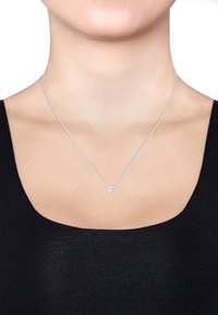 Silberne Kettenhalskette mit einem kleinen, eleganten Buchstaben "F" Anhänger, präsentiert auf einem schwarzen Oberteil vor einem schlichten weißen Hintergrund.