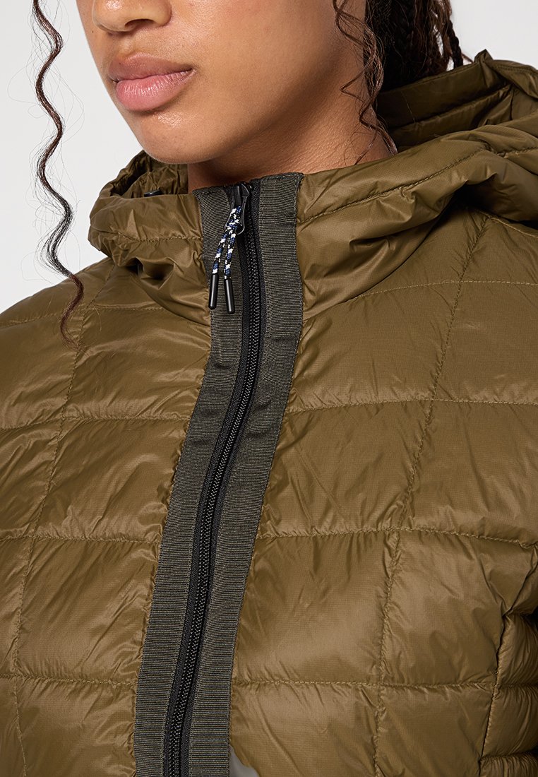 Nahaufnahme einer Person in einer olivgrünen, gesteppten Jacke mit hohem Kragen, schwarzem Reißverschluss und Kapuze, die einen Teil des Gesichts und der Haare zeigt.