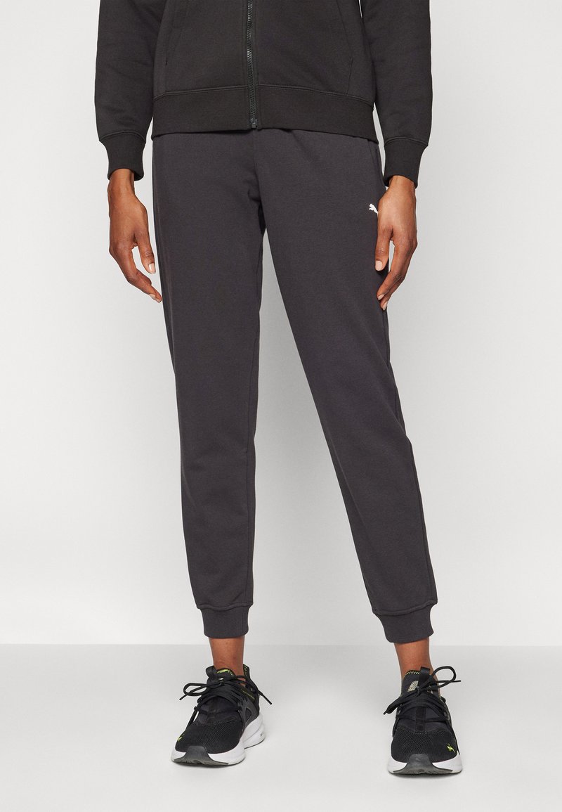 Graue Sweatpants mit elastischen Bündchen, aus weichem Material gefertigt. Mit einem kleinen weißen Logo auf der linken Seite und getragen mit schwarzen Sneakers.